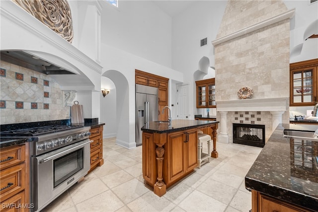 kitchen featuring a kitchen island with sink, a tiled fireplace, high quality appliances, sink, and a towering ceiling