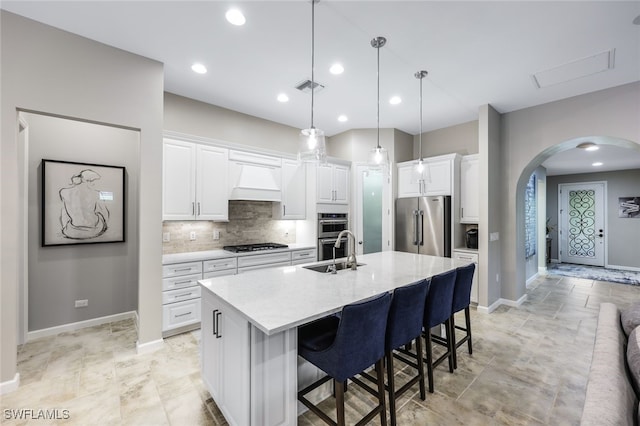 kitchen featuring premium range hood, a kitchen island with sink, white cabinetry, stainless steel appliances, and decorative light fixtures