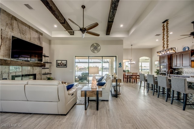 living room with beam ceiling, ceiling fan with notable chandelier, a fireplace, and light wood-type flooring