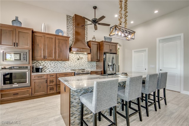 kitchen featuring light stone countertops, appliances with stainless steel finishes, custom range hood, a breakfast bar area, and a center island with sink