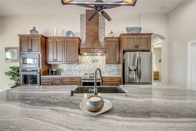 kitchen with appliances with stainless steel finishes, backsplash, ceiling fan, light stone counters, and premium range hood