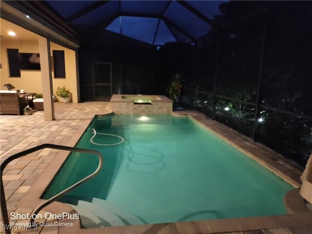 pool at night featuring glass enclosure, a hot tub, and a patio area