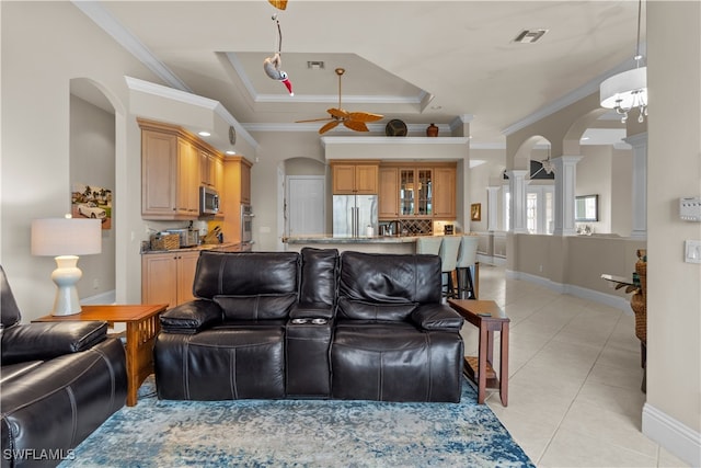 living room featuring ceiling fan with notable chandelier, a raised ceiling, ornamental molding, light tile patterned flooring, and decorative columns