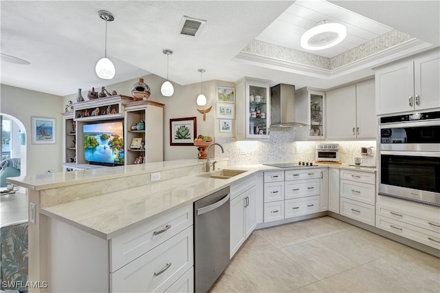 kitchen with wall chimney range hood, sink, a raised ceiling, kitchen peninsula, and stainless steel appliances