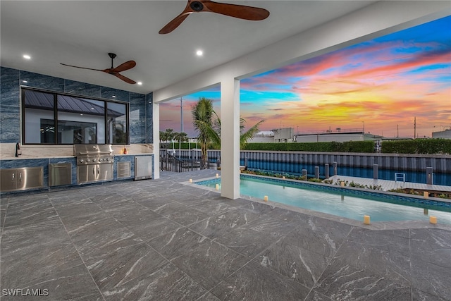 pool at dusk with grilling area, a patio area, ceiling fan, and exterior kitchen