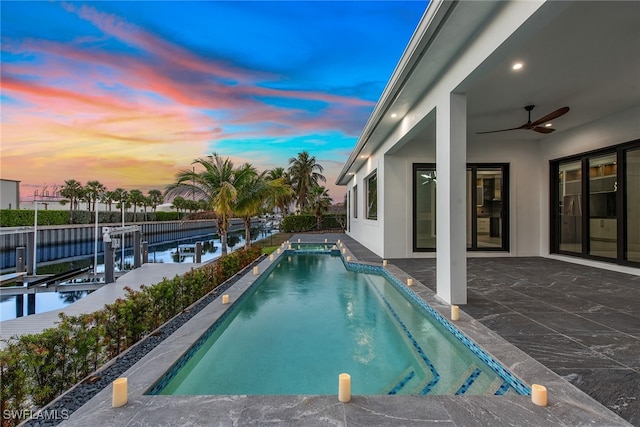 pool at dusk with ceiling fan, a water view, a patio, and a boat dock