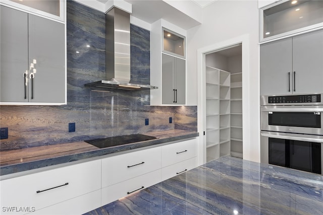 kitchen with decorative backsplash, dark stone counters, wall chimney exhaust hood, stainless steel double oven, and black electric cooktop