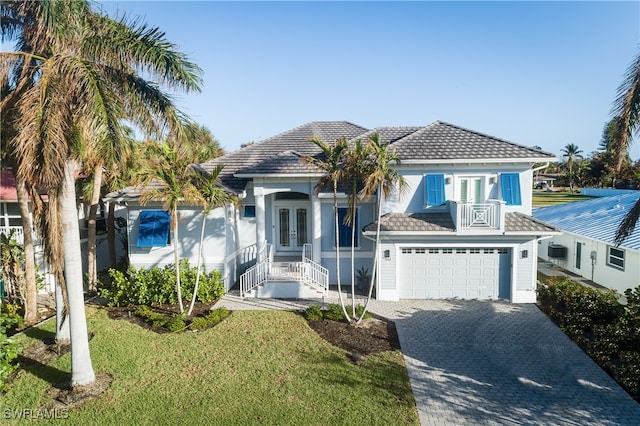 view of front of home with an attached garage, a balcony, french doors, decorative driveway, and a front lawn