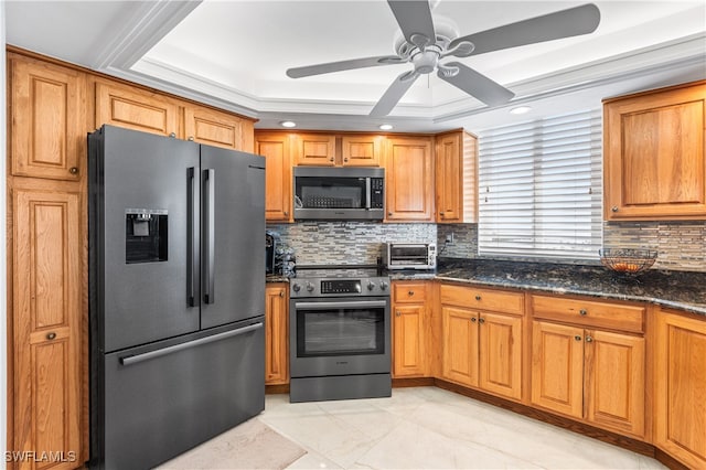 kitchen featuring appliances with stainless steel finishes, dark stone counters, ceiling fan, a tray ceiling, and decorative backsplash