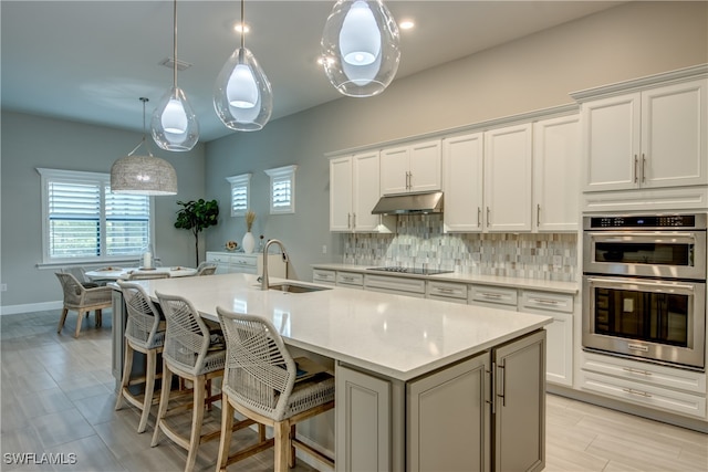 kitchen with stainless steel double oven, a center island with sink, sink, decorative light fixtures, and white cabinets