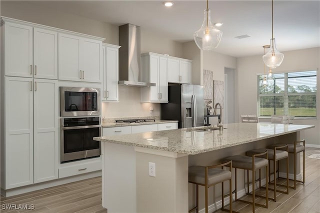 kitchen featuring hanging light fixtures, appliances with stainless steel finishes, an island with sink, white cabinets, and wall chimney exhaust hood