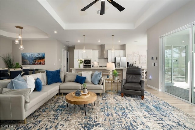 living room with light wood finished floors, baseboards, recessed lighting, a raised ceiling, and a ceiling fan