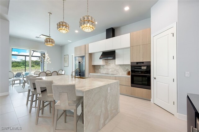 kitchen with light stone counters, pendant lighting, an island with sink, a chandelier, and wall chimney exhaust hood