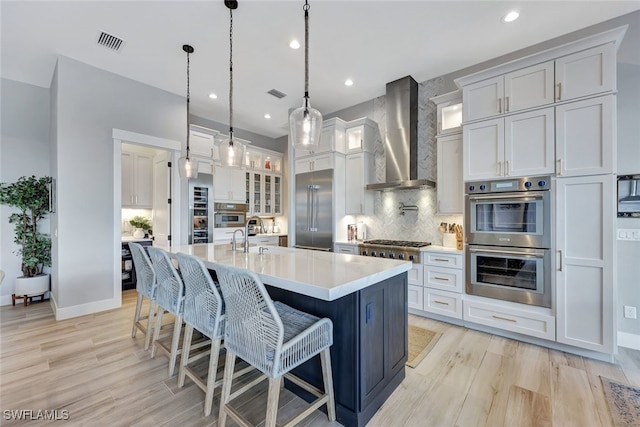 kitchen with a kitchen island with sink, wall chimney exhaust hood, decorative light fixtures, light hardwood / wood-style floors, and stainless steel appliances