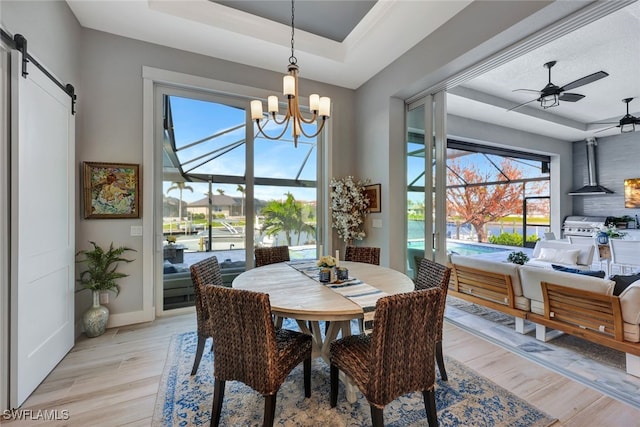 dining space with a barn door, a tray ceiling, a water view, ceiling fan with notable chandelier, and light wood-type flooring