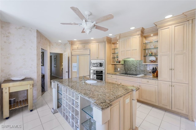 kitchen featuring built in appliances, light stone counters, light brown cabinets, ceiling fan, and a kitchen island with sink