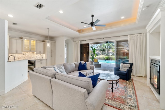 living room with beverage cooler, light tile patterned floors, ornamental molding, ceiling fan, and a raised ceiling