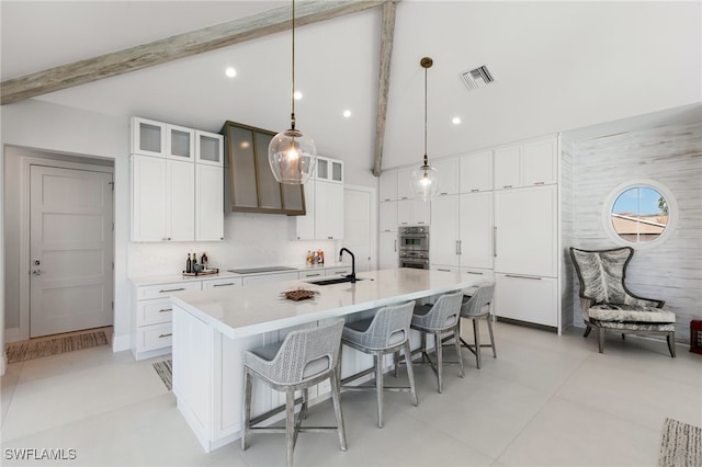 kitchen with sink, hanging light fixtures, beamed ceiling, an island with sink, and white cabinets