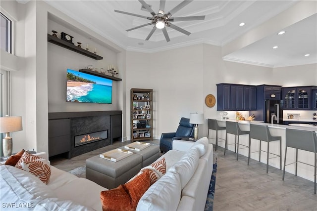 living room with ceiling fan, light wood-style flooring, a towering ceiling, a glass covered fireplace, and crown molding