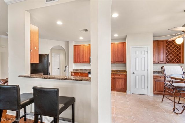 kitchen with black fridge, kitchen peninsula, light tile patterned floors, a breakfast bar area, and dark stone countertops