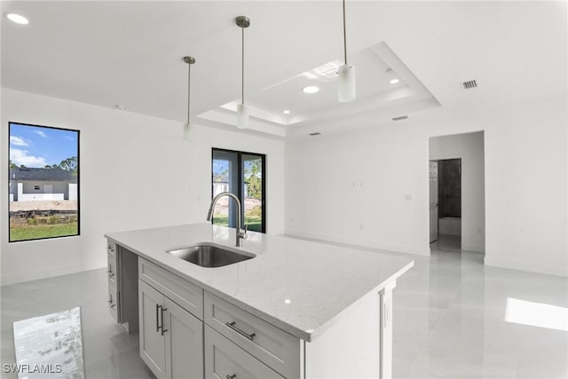 kitchen featuring light stone counters, a raised ceiling, sink, pendant lighting, and an island with sink