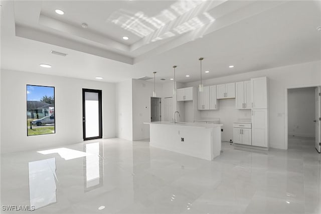 kitchen featuring a raised ceiling, decorative light fixtures, white cabinetry, and an island with sink