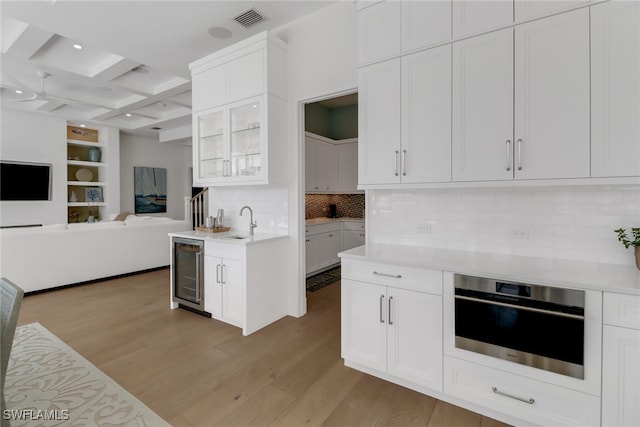 kitchen featuring tasteful backsplash, beam ceiling, beverage cooler, stainless steel oven, and light wood-type flooring