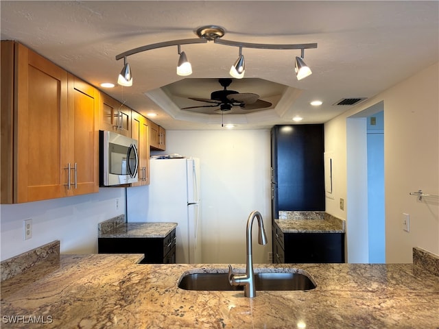 kitchen with ceiling fan, sink, light stone counters, white fridge, and a tray ceiling