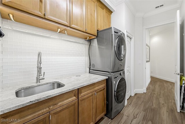 clothes washing area featuring sink, cabinets, ornamental molding, stacked washer and clothes dryer, and light wood-type flooring