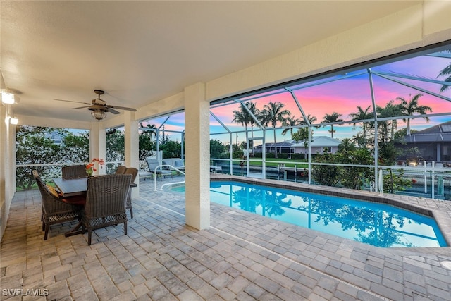 pool at dusk with glass enclosure, a patio, and ceiling fan