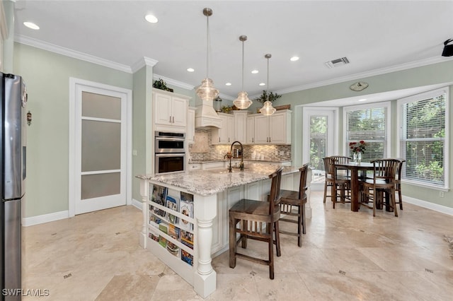 kitchen with light stone countertops, stainless steel appliances, backsplash, decorative light fixtures, and a kitchen island with sink