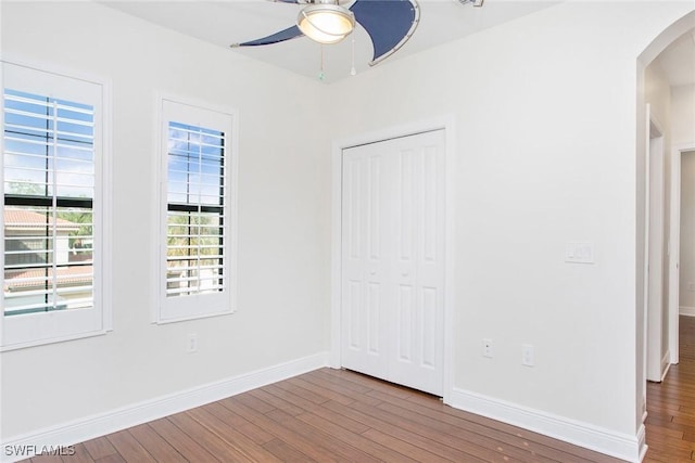 unfurnished bedroom with arched walkways, a closet, a ceiling fan, wood finished floors, and baseboards