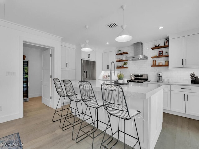 kitchen with appliances with stainless steel finishes, white cabinetry, hanging light fixtures, a center island with sink, and wall chimney exhaust hood