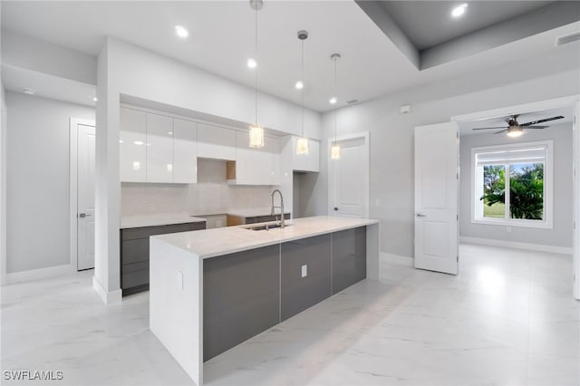 kitchen with ceiling fan, sink, white cabinetry, hanging light fixtures, and an island with sink