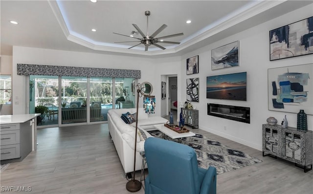 living room featuring ceiling fan, ornamental molding, light wood-type flooring, a tray ceiling, and a glass covered fireplace