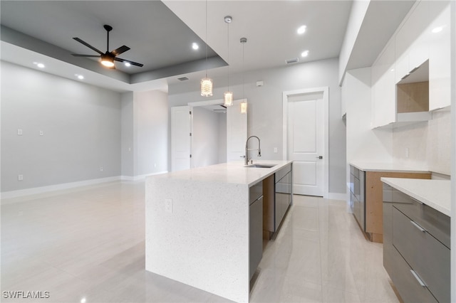 kitchen with a tray ceiling, ceiling fan, sink, decorative light fixtures, and an island with sink