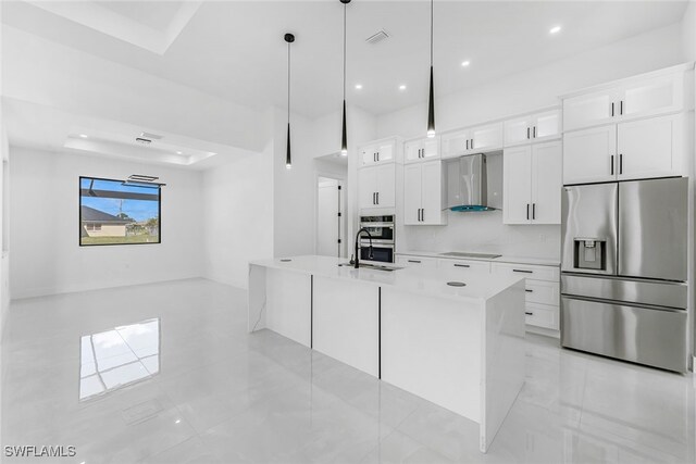 kitchen featuring appliances with stainless steel finishes, a kitchen island with sink, wall chimney range hood, pendant lighting, and white cabinetry