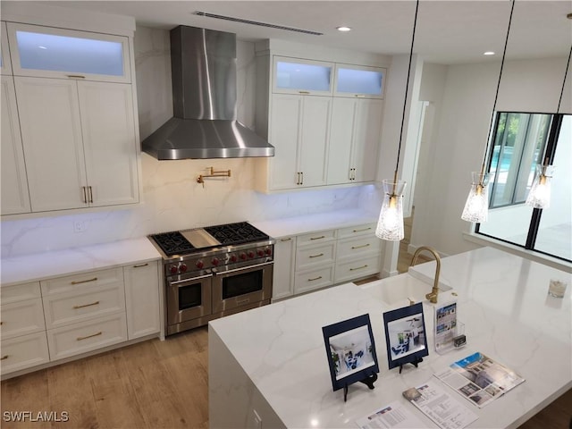 kitchen with light wood-type flooring, wall chimney range hood, light stone countertops, range with two ovens, and white cabinets