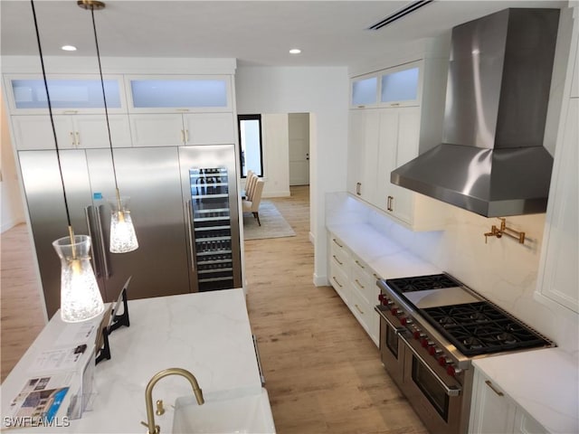 kitchen with wall chimney range hood, white cabinetry, hanging light fixtures, light stone counters, and premium appliances