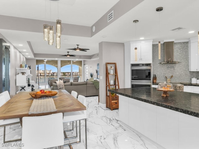 kitchen with pendant lighting, wall chimney exhaust hood, double oven, backsplash, and white cabinetry