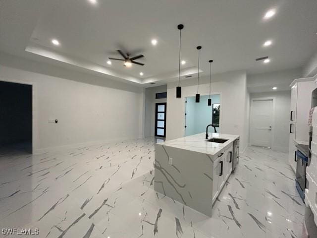 kitchen with a kitchen island with sink, sink, hanging light fixtures, light stone counters, and white cabinetry