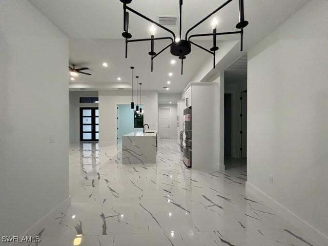 kitchen with light stone counters, ceiling fan with notable chandelier, decorative light fixtures, white cabinets, and an island with sink