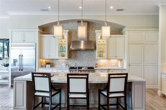 kitchen featuring a center island with sink, hanging light fixtures, ornamental molding, stainless steel built in fridge, and wall chimney exhaust hood