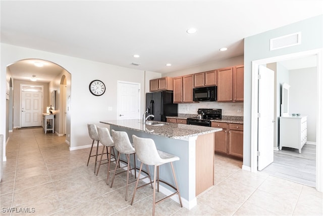 kitchen with a breakfast bar area, light stone countertops, black appliances, an island with sink, and light tile patterned flooring