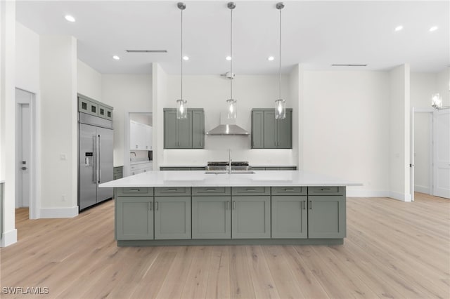 kitchen with stainless steel built in refrigerator, wall chimney exhaust hood, an island with sink, light wood-type flooring, and decorative light fixtures