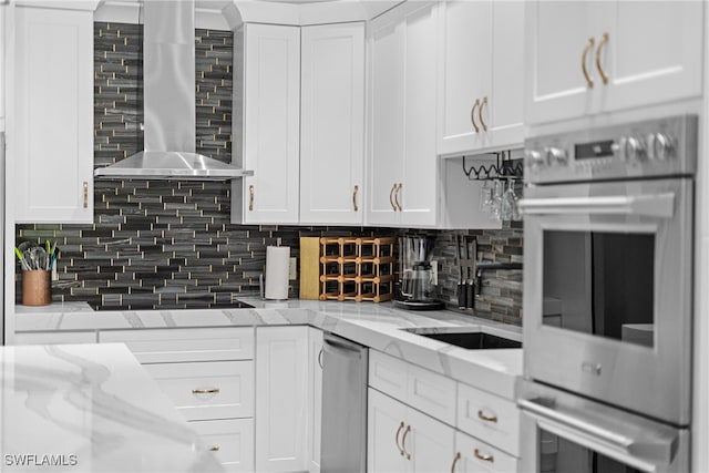 kitchen featuring white cabinetry, wall chimney range hood, stainless steel double oven, and light stone counters