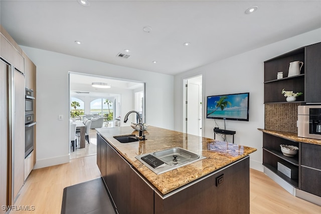 kitchen featuring a center island with sink, sink, light stone counters, dark brown cabinets, and light wood-type flooring