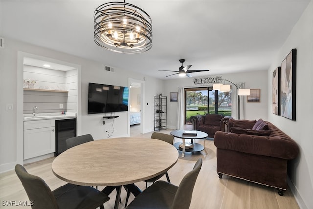 dining space with beverage cooler, ceiling fan with notable chandelier, sink, and light wood-type flooring