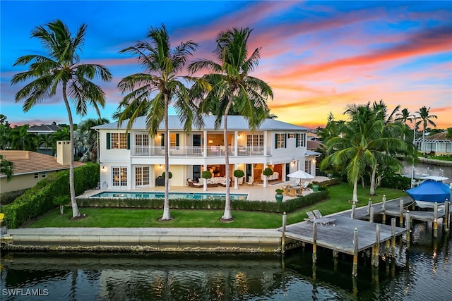 back house at dusk with a patio area, a water view, a lawn, and a balcony