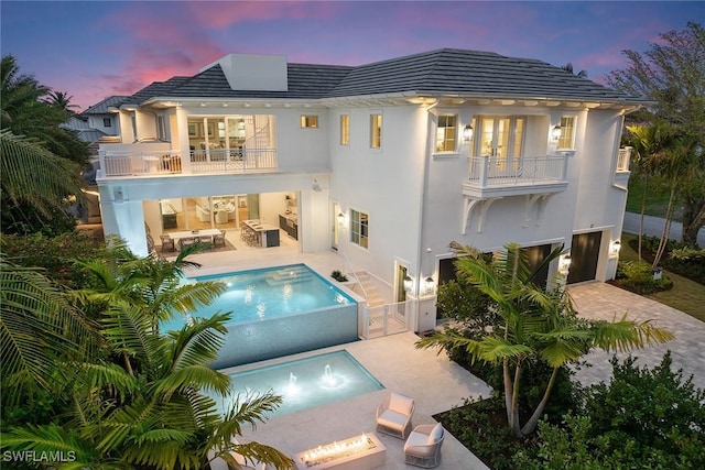 back house at dusk with an outdoor living space, a pool with hot tub, a balcony, and a patio